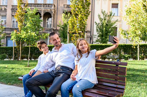 Young Handsome Man is Enjoying with Children on Summer Day in a Park While Sitting on a Bench and Embracing his Kids.