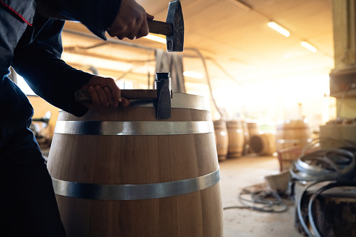 At the wood workshop, a Caucasian male Cooper using hammer to adjust the metal hoop on oak barrel