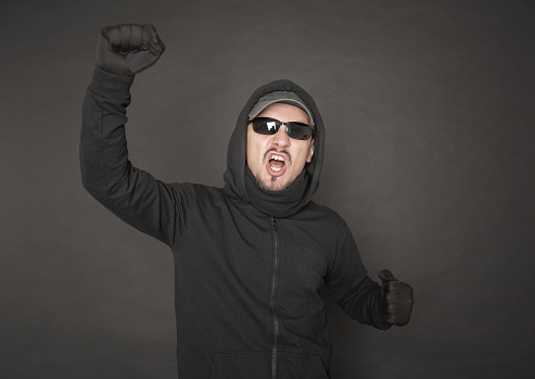 Screaming Man in the black hoody with hood wearing sunglasses with fist up on dark background