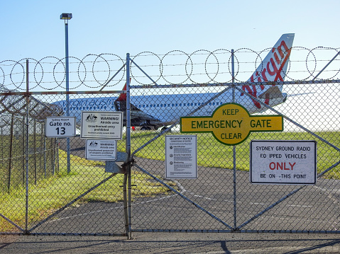 Gate 13 of Sydney Kingsford-Smith Airport with warning signs and barbed wire.  In the background is a Virgin Australia Boeing 737 taxiing to the runway.  This image was taken from near Shep's Tower Mound, a public viewing area off Ross Smith Avenue, on a sunny afternoon on 4 February 2023.