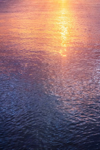 The serene beauty of a river at sunset. The golden sunlight reflects on the water, creating a sparkling effect that showcases a variety of colors such as orange, purple, and blue. The intricate ripples on the surface of the river add depth and texture to the scene, making it a mesmerizing and captivating view.