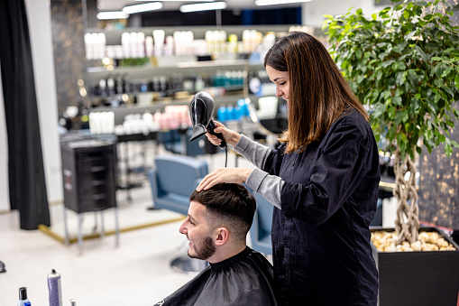 A female worker in a barbershop is using a hairdryer after haircutting a young male customer.