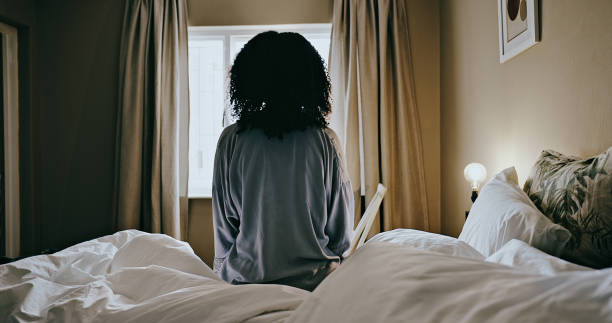 le matin, déprimée et la femme se réveillent en pensant sur son lit dans sa chambre en regardant par la fenêtre et assise. lever du soleil, vue solitaire et arrière d’une dame se relaxant en réfléchissant aux souvenirs de sa maison - solitaire bird photos et images de collection