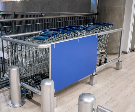 Row of shopping cart with blue handle lined up on wood floor by barrier wall at parking lot of supermarket.