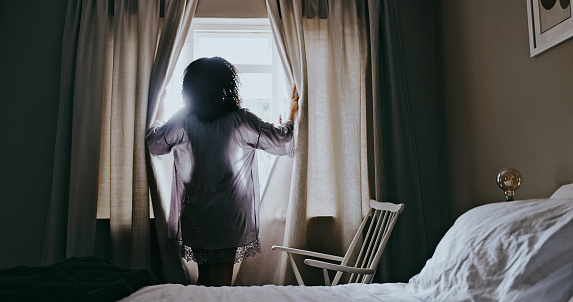 Bedroom, window and woman opening curtains ready for morning and looking at the sunrise. Rear view, wake up and lady relaxing in her home or house after sleep, rest and enjoying the sunlight