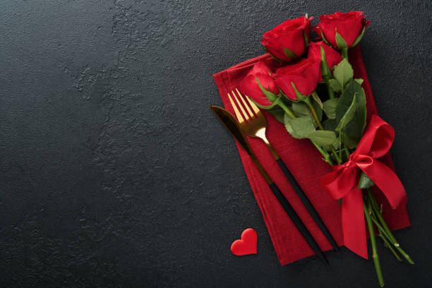 istock Valentines day romantic table setting. Empty closeup of red roses, wine, candles, dinner black plate, knife, fork and decorative silk hearts on black background. Holiday concept. Copy space. Top view 1462678295
