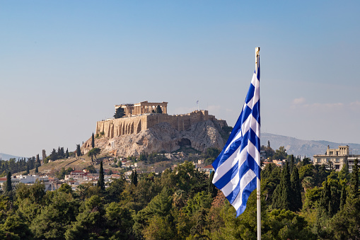 A picture of the Acropolis of Athens, and the Parthenon, as seen above a tree line and the Greek flag in the foreground.
