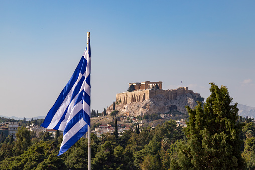 A picture of the Acropolis of Athens, and the Parthenon, as seen above a tree line and the Greek flag in the foreground.