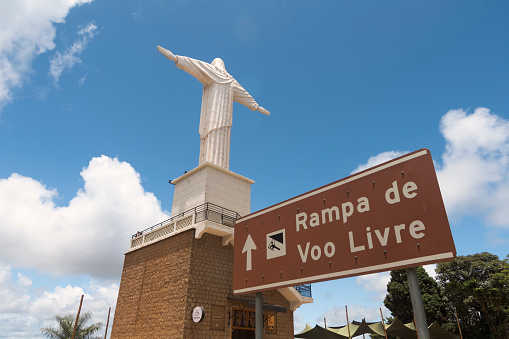 Poços de Caldas, Minas Gerais, Brazil. January 27, 2023. Sign indicating access to free flight ramp. Statue of Christ. Blue sky with white clouds.