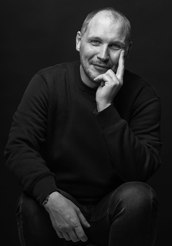 Lifestyle concept. Studio portrait of happy man with sweatshirt looking at camera with smile and holding one hand on cheek. Black studio background. Black and white image