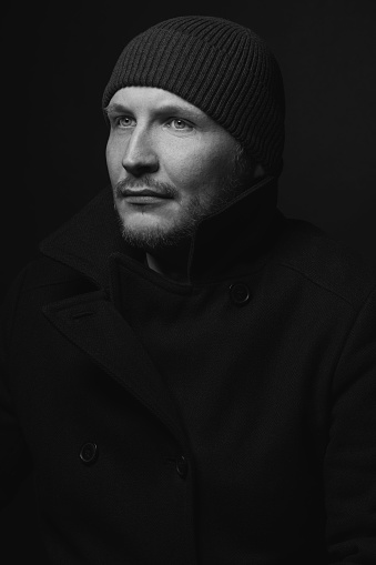 Lifestyle, fashion, occupation concept. Man with winter hat and black coat studio portrait. Model with beard looking aside the camera with serious look. Dark studio background. Black and white image