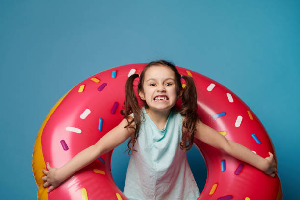 bambina maliziosa che fa facce mentre posa all'interno di un tubo da nuoto rosa, su sfondo isolato di colore blu - swimming tube inflatable circle foto e immagini stock