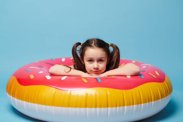 little child girl in swimsuit, inside a swim ring - a colorful pink donut, smiling at camera, isolated blue background. - swimming tube inflatable circle imagens e fotografias de stock