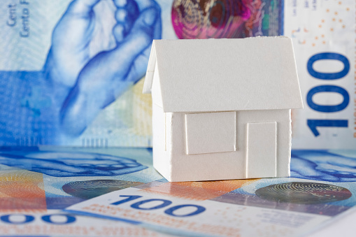 A model of a detached house made of white paper stands on Swiss banknotes and is visible against the background of the same banknotes, which could mean real estate, mortgage or just a paper toy or other real estate meaning