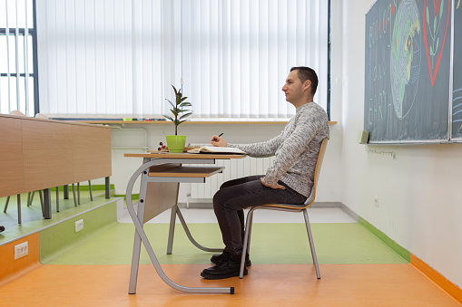 A side view of one young male Caucasian school teacher sitting at the desk in the classroom and giving a lesson about ecology and planet Earth.