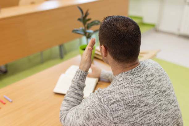 A rear view of one young male Caucasian school teacher sitting at the desk in the classroom.