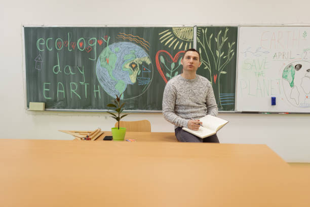 A front view of one Caucasian male teacher sitting on the desk in the classroom and holding a notebook and a pen.