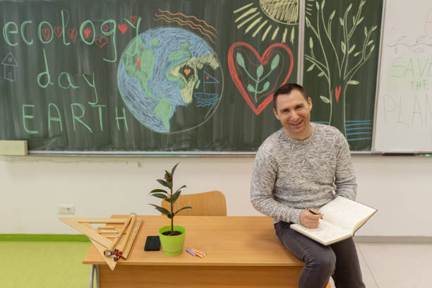 One cheerful Caucasian male professor sitting on the desk in the classroom and holding a notebook and a pen, checking who is absent.  Save the planet Earth concept.