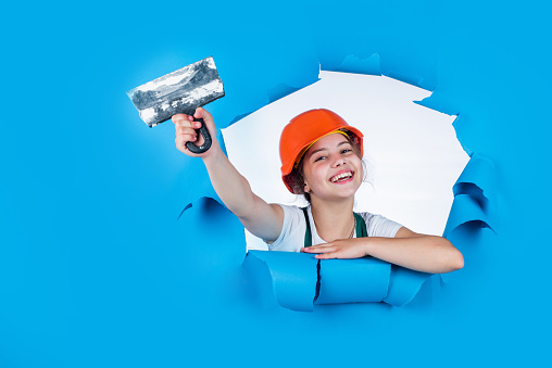 builder in coveralls and helmet with spatula. kid repair wall with spackling paste. Home renovation. Kid with tool. happy small construction girl with spatula. teen girl use putty knife.