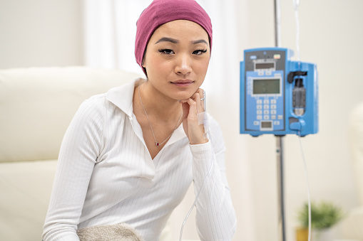 A young woman of Asian decent, sits in a treatment clinic as she receives her Chemotherapy treatment.  She is dressed comfortably with a headscarf and blanket on to keep warm as her treatment flows intravenously.