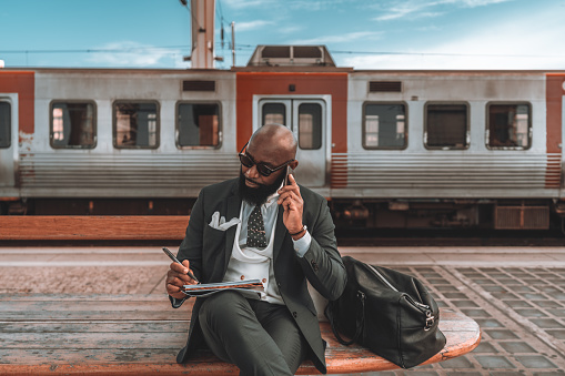 A black man multitasks on a blue-sky day, sitting on a wooden park bench while talking on the phone and jotting down notes. His weekender sits by his side, as a train buzzes by in the background.
