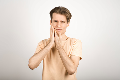 A young Caucasian blond guy with a stylish hairstyle presses his hand on his cheek because of a toothache isolated on a white studio background. The concept of medicine and health.