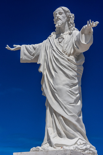 closeup of a stone Jesus statue in a cemetery, with arms outstretched backed by a bright blue sky