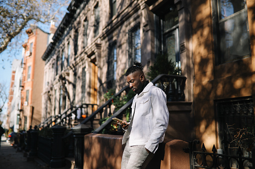 Lifestyle portrait of a fashionable, young black millennial man in Brooklyn, Park Slope district, NYC. He is wearing a white jacket, checking apps on the smartphone.
