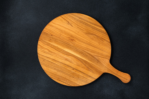 Round empty wooden charcuterie serving board with handle on old black concrete background, shabby table. Top view. Food template, copy space. Flat lay design, mockup. Layout, cooking concept, frame