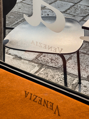 Travel destination Venice, Italy concept. The shadow of letters on a display window at a bar are reflecting upside down on an orange sofa surface. Sunlight falls indoors. Outdoors a part of a chair stand on cobblestone. Poster template background, copy space.