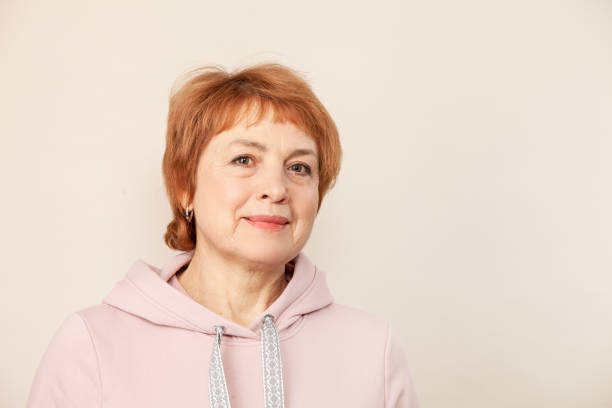 Studio portrait of a white active senior woman with red hair in a pink hooded shirt against a beige background stock photo