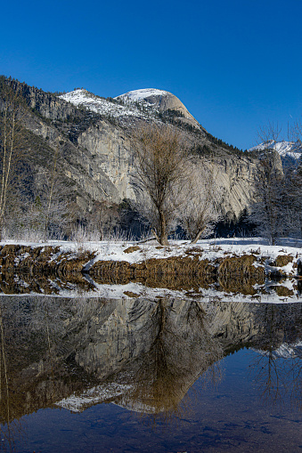 In California, Yosemite National Park is a US national park bordered to the southeast by the Sierra National Forest and to the northwest by the Stanislaus National Forest. On a sunny morning, the sky was clear and cloudless. Reflections of distant mountains could be seen in the still, frozen water. A few barren trees can be found nearby.