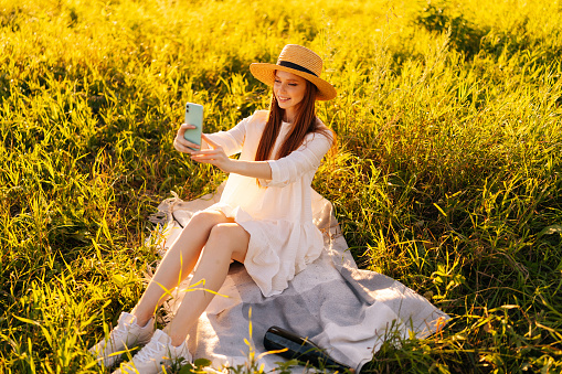 Smiling red-haired young female tourist in straw hat and white dress using taking selfie shot on smartphone looking to mobile screen, sitting on beautiful field of green grass on summer sunny day