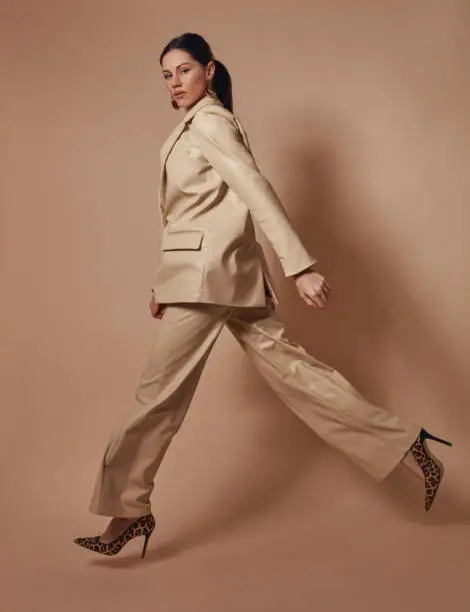 Masculine-looking beautiful woman in a leather suit is jumping in front of a brown background.