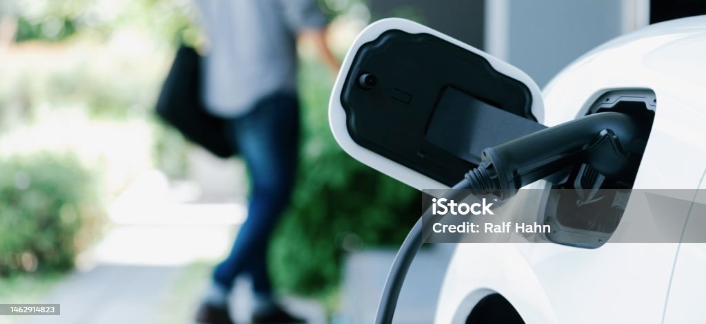 Focus EV charging station at home with blur progressive man in background. Focus electric car charging at home charging station with blurred progressive man walking in the background. Electric car using renewable clean for eco-friendly concept. Car Stock Photo