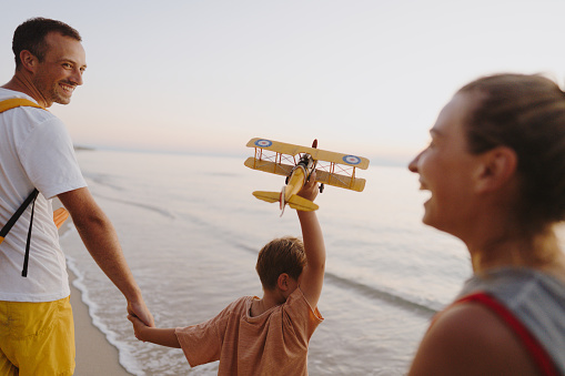 Photo of a happy, smiling family enjoy evening walk down the beach, while little boy is playing with airplane toy