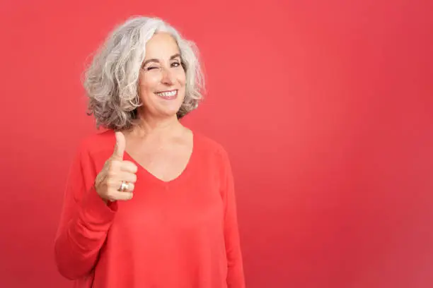 Studio portrait with red background of a mature woman winking as she gives a thumbs up in approval