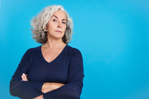 Studio portrait with blue background of a mature woman with grey hair looking at camera with a distrustful expression