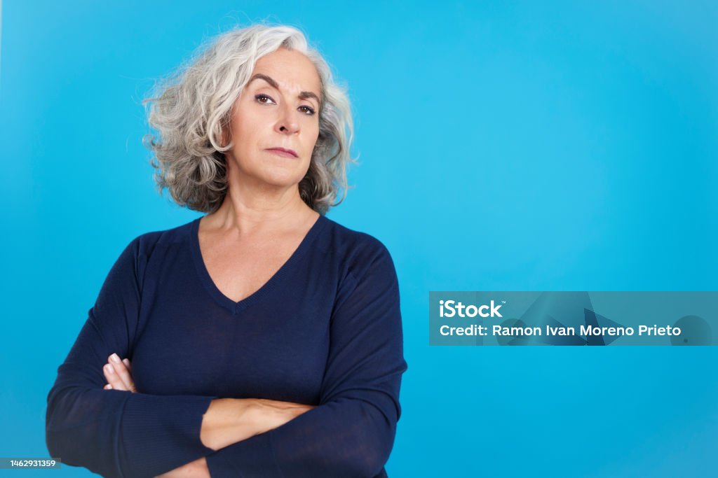 Mature woman looking at camera with a distrustful expression Studio portrait with blue background of a mature woman with grey hair looking at camera with a distrustful expression Suspicion Stock Photo