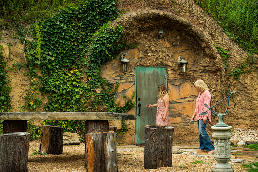Blond women approach the hobbit-like earthen salt cave beside a rustic table with tree stump stools outside the Pomona Salt Cave and Spa, West Virginia, USA