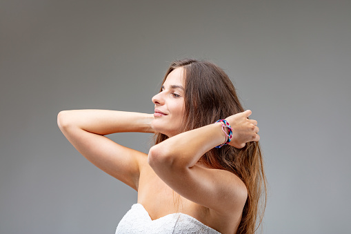 Three-quarter portrait of young woman serenely stroking her hair. They are long and brown, and she is wearing a white dress that leaves her chest and shoulders uncovered, and the only color besides he