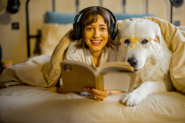 Young woman with her cute dog in eyeglasses reading a book while lying togther under a blanket in bedroom. Spending leisure time, friendship with pet concept