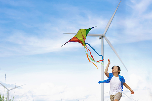 Progressive happy carefree boy concept. Young boy running along the road and flying kite at natural scenic on countryside, clear sky and sunny day with mountain and wind turbine background