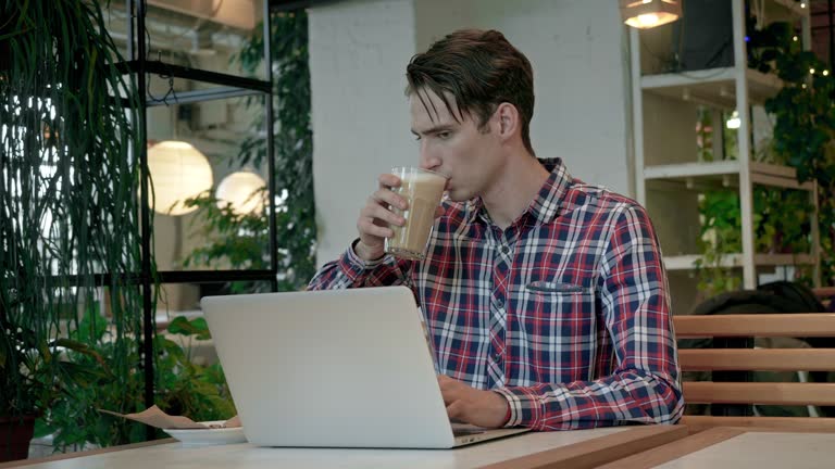 Young man sipping coffee and typing on a laptop in a coffee shop