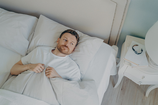 View from above of stressed young caucasian male lying in bed with his eyes open and being unable to sleep, man feeling unhappy and tired from insomnia problems. Bedtime and rest trouble concept