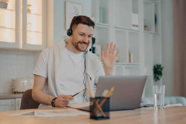 istock E learning concept. Happy bearded man student has video conference studies online waves palm in laptop display greets teacher works distantly uses headset makes notes in diary poses over home interior 1462977048