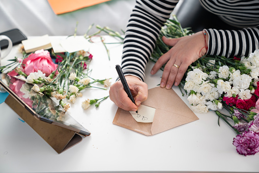 Close up photo of mature woman hands arranging carnation flower bouquet in living room for selling online. She is sitting on sofa next to window, writing a message on envelope and having video call on digital tablet. Shot under daylight.