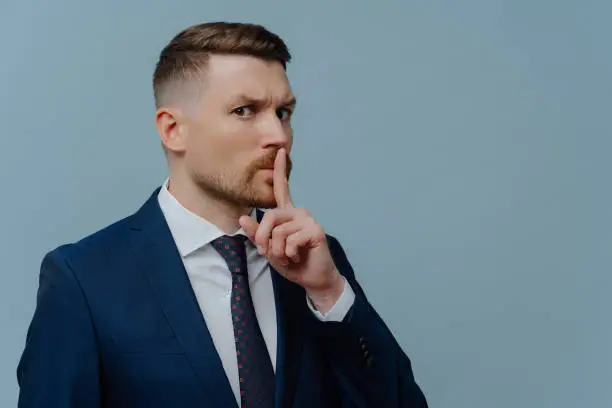Serious businessman in suit asking to keep private information on secret and showing shh sign while standing against light steel blue background, guy looking at camera and keeping finger near mouth