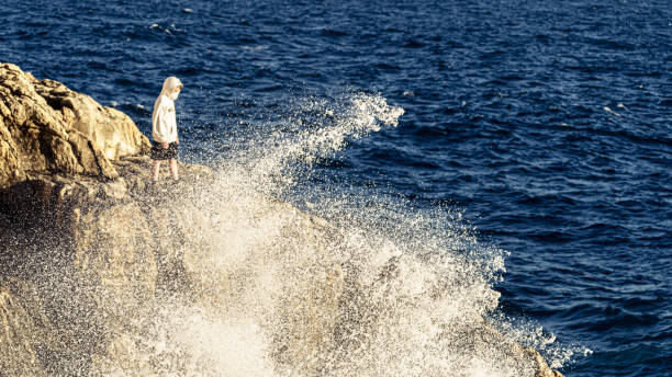 A boy standing on a cliff next to a crushing wave.