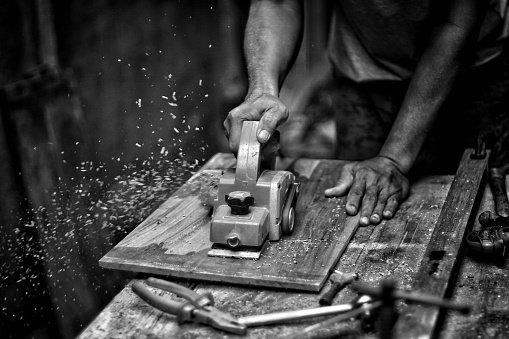 A grayscale shot of a carpenter working with a plane on the wood